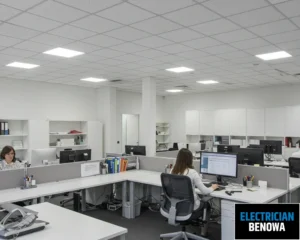 An office with desks and people working, illuminated by bright ceiling lights.